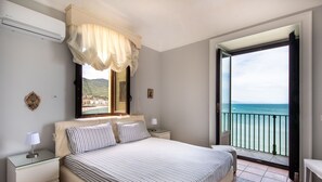Ein Schlafzimmer mit einem großen Bett, einem Nachttisch, einer Lampe und Meerblick durch eine geöffnete Tür.