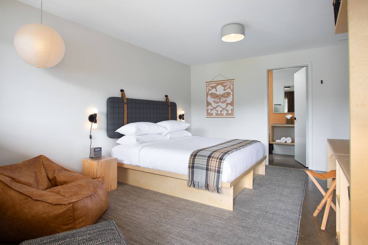 Um quarto moderno com cama, mesas de cabeceira, um banquinho de madeira, uma poltrona de couro marrom, um tapete cinza e uma escrivaninha de madeira.