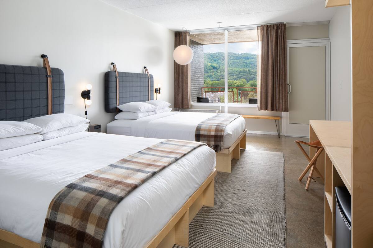 Quarto de hotel com duas camas, uma escrivaninha, uma cadeira, uma janela grande com cortinas e vista para vegetação.