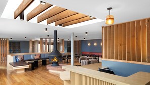 A modern interior with a wooden reception desk, a seating area with a fire feature, and a high ceiling with exposed beams.