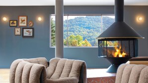 A room with a fireplace, two chairs, a window with a view of a forest, and framed pictures on the wall.