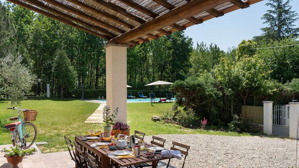 En udendørs terrasse med et træbord dækket til spisning, omgivet af stole, og en pergola over hovedet.