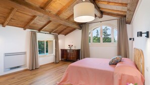 Et soveværelse med træloft, en seng med pink dyne, et træmøbel og et vindue med udsigt til træer, samt en loftlampe.