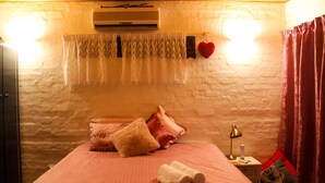 Une chambre à coucher avec un lit, des oreillers, une table de chevet avec une lampe, un climatiseur fixé au mur et une fenêtre avec des rideaux.