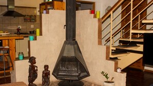 Une cuisine avec un poêle central, un coin repas comprenant une table et des chaises, et un escalier menant à l’étage supérieur.