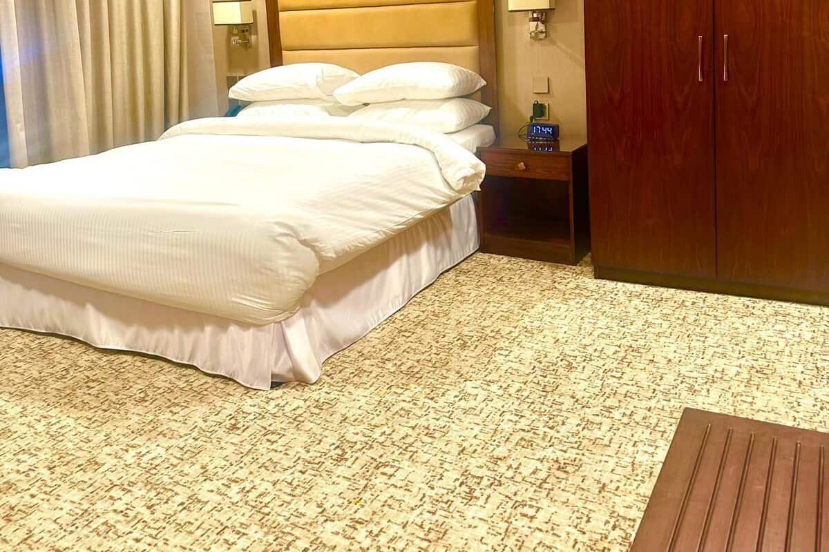 A hotel room with a large bed, a wooden wardrobe, a nightstand with a lamp, and a floral artwork on the wall.