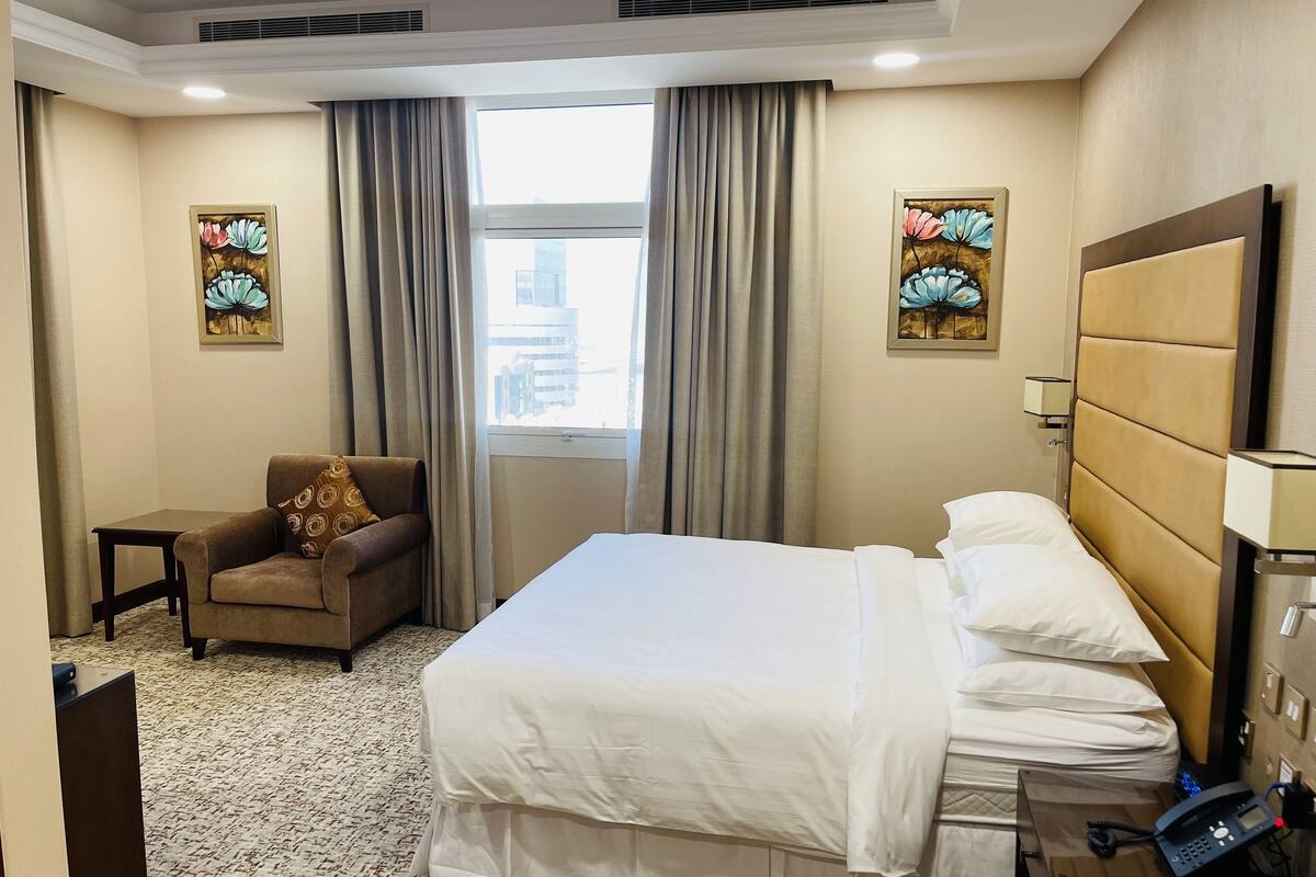 A hotel room with a bed, a chair, a desk, and two paintings on the walls.