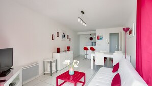 Una sala de estar moderna con un sofá blanco, detalles en rojo, un televisor y una zona de comedor con sillas rojas.
