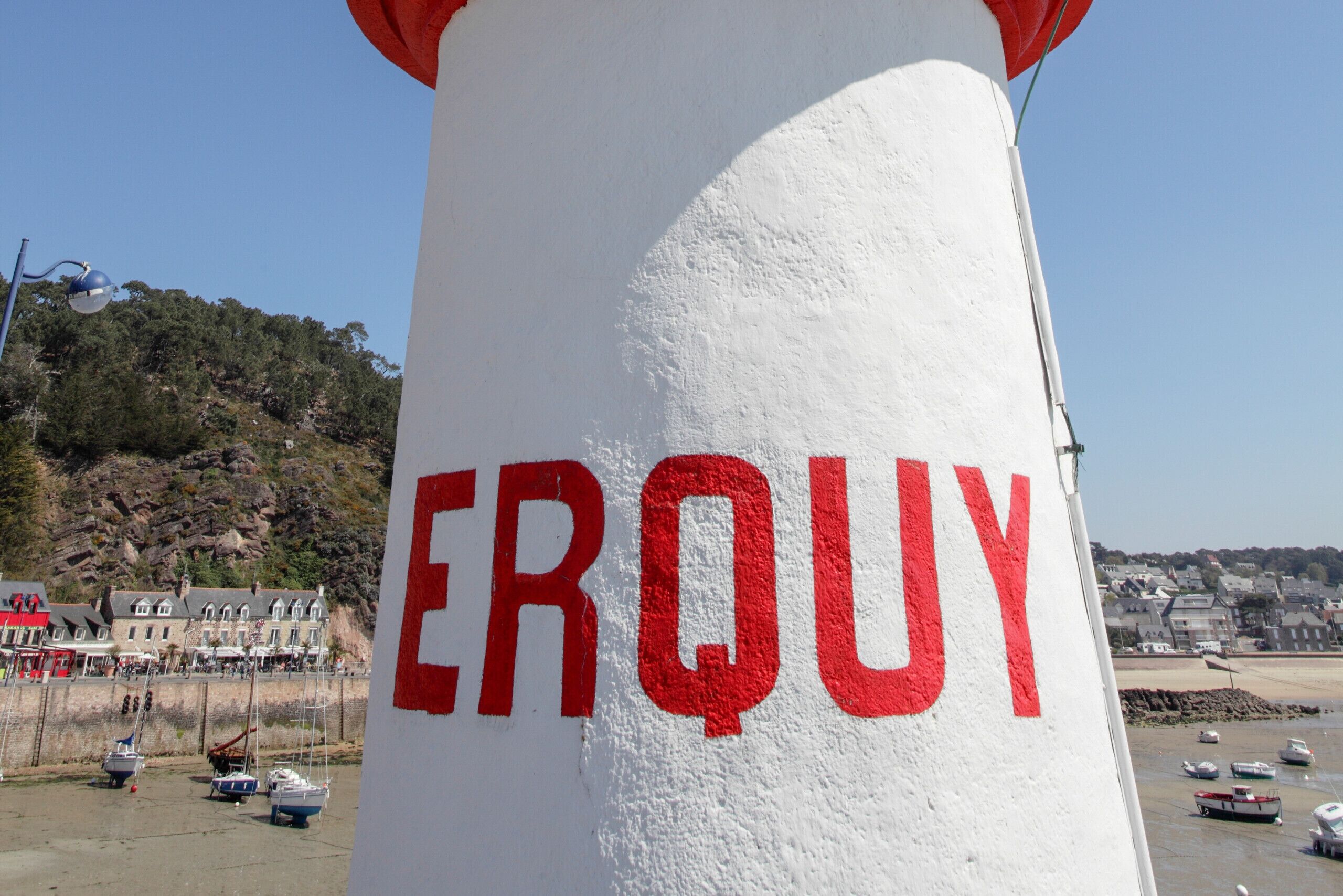 Ein Leuchtturm mit der Aufschrift 'ERQUY', umgeben von Booten und einer Küstenstadt im Hintergrund.
