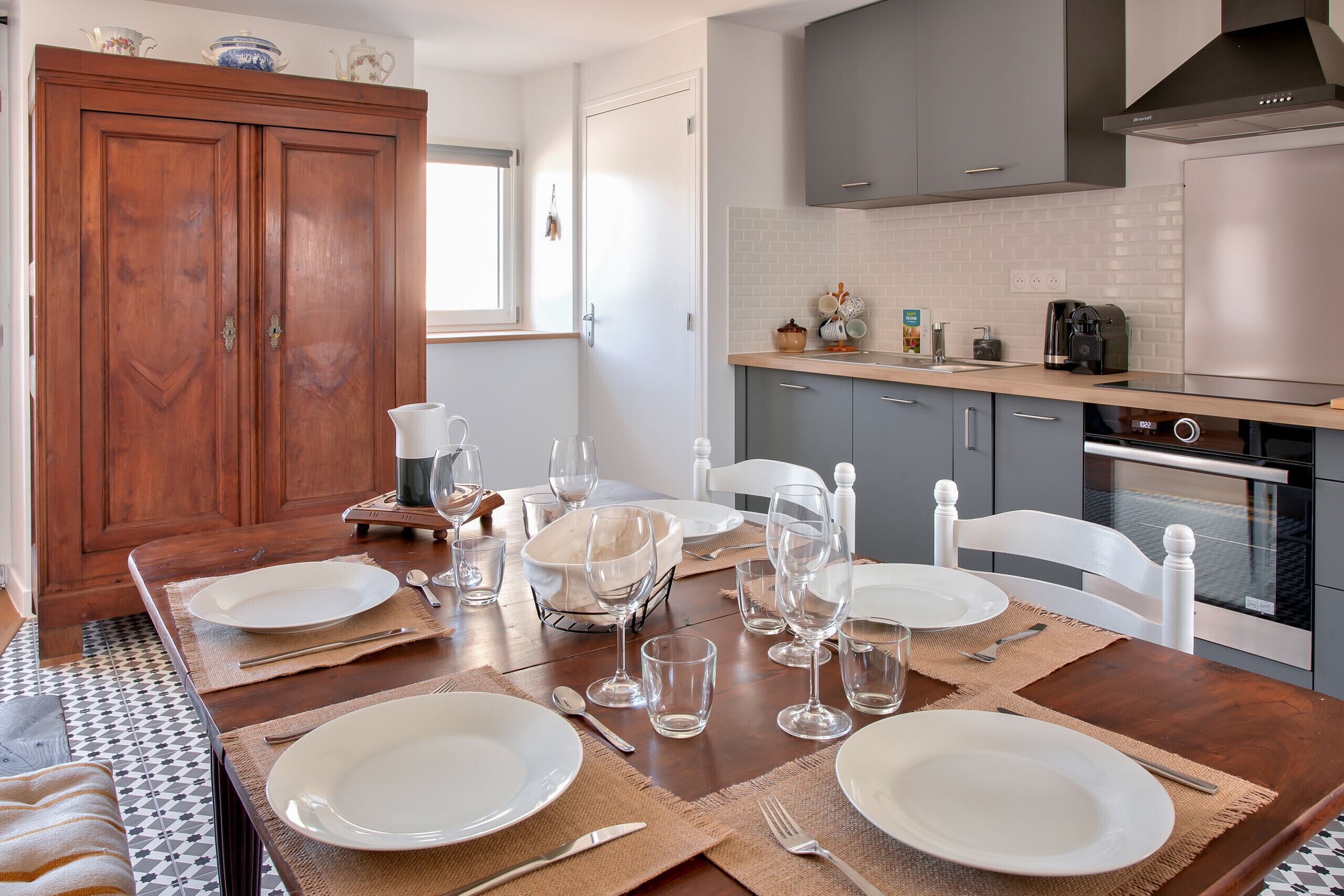 A dining area with a wooden table set for six, surrounded by grey cabinets and a modern kitchen in the background.