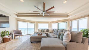 A spacious living room with a large sectional sofa, a ceiling fan, and large windows offering a view of the ocean. - Stunning Oceanfront Home w/ Panoramic Views (Oceanside)
