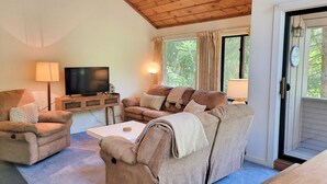 A cozy living room with a wooden ceiling, a television on a wooden stand, a sofa, a coffee table, and a door leading outside. - Open concept, deck, fireplace, AC, pet-friendly, near Loon. C6301 by LRS (Lincoln)