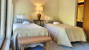 A bedroom with two beds, a bedside table with a lamp, a bench, and a door leading to a bathroom. - Open concept, deck, fireplace, AC, pet-friendly, near Loon. C6301 by LRS (Lincoln)