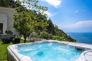 A hot tub with a view of the sea, surrounded by greenery and a building with outdoor seating.