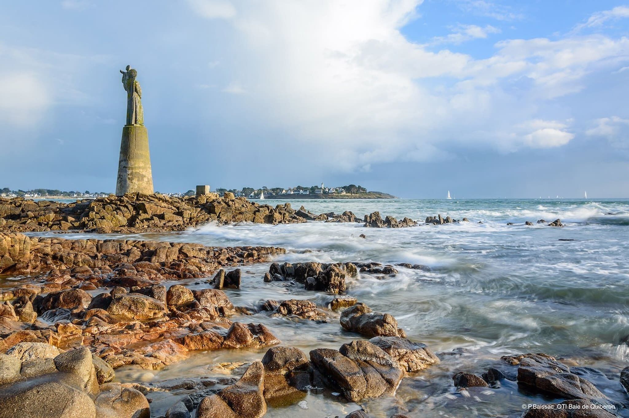 Una costa rocciosa con una statua in evidenza su un piedistallo, un faro sullo sfondo e una località costiera dall'altra parte dell'acqua.