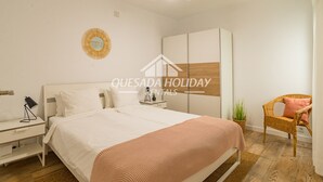 Um quarto com cama, mesas de cabeceira, guarda-roupa, ventoinha no teto e uma cadeira de vime.
