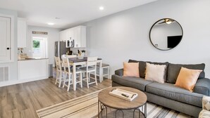 Eine moderne Küche mit weißen Schränken, ein Essbereich mit weißen Stühlen, ein graues Sofa mit Kissen und ein runder Spiegel an der Wand.