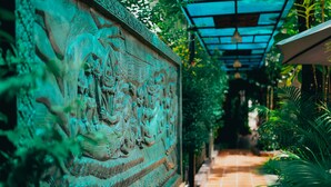 A corridor with a detailed stone wall featuring carvings, surrounded by lush greenery and a tiled floor. - Sophavady D'Angkor (Siem Reap)