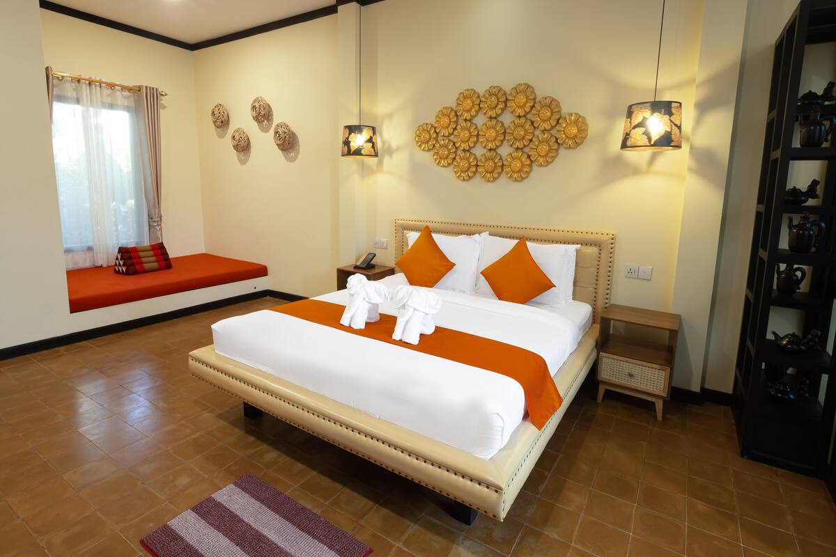 A well-lit bedroom with a large bed, a bedside table, a nightstand, a shelf with decorative items, and a window with curtains.