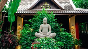 A statue of a seated figure in a meditative pose, surrounded by lush greenery and a traditional building with a tiled roof. - Sophavady D'Angkor (Siem Reap)