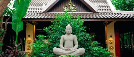 Una estatua de una figura sentada en posición meditativa, rodeada de abundante vegetación y un edificio tradicional con tejado de baldosas.