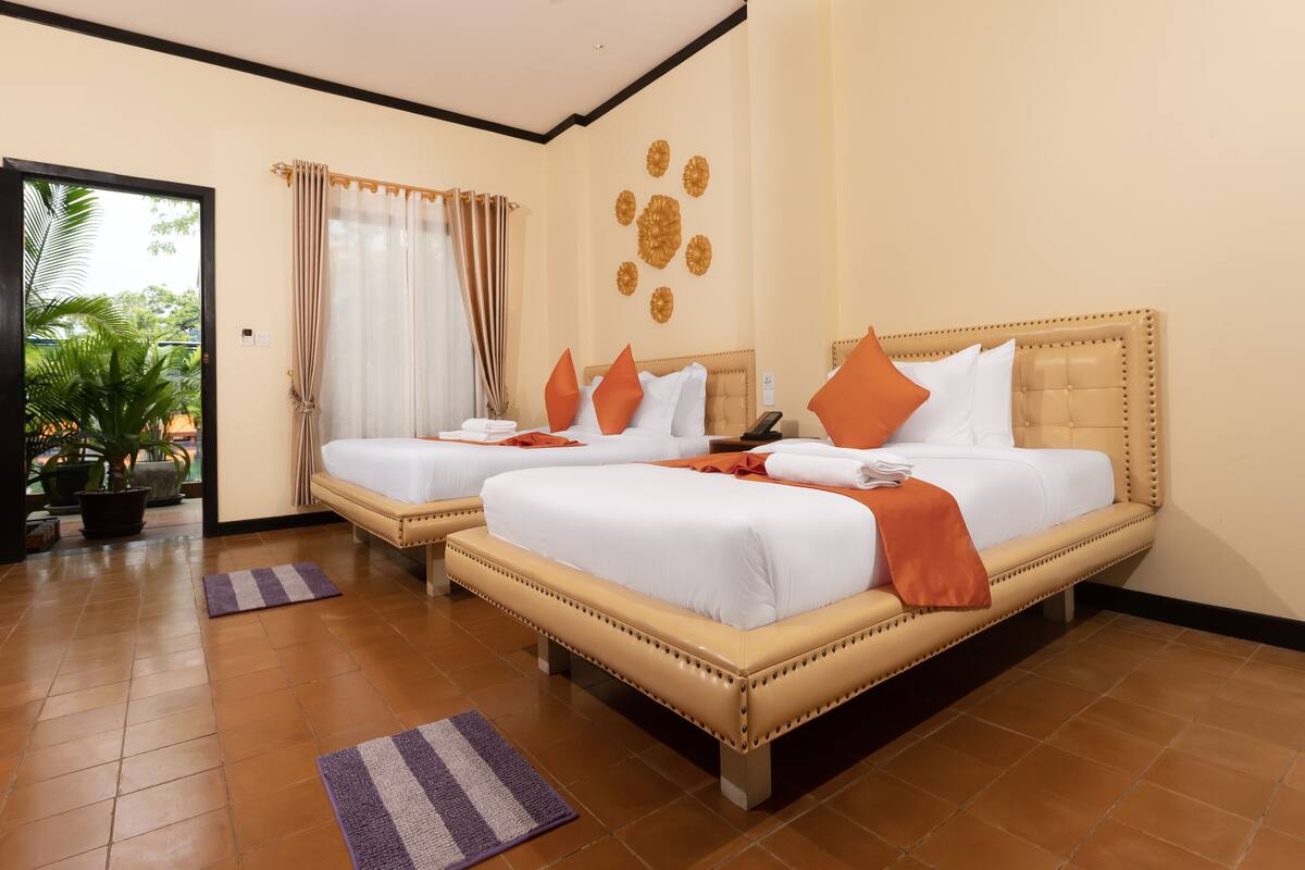 A hotel room with two beds, a large potted plant, a view of the outdoors, and a ceiling fan.