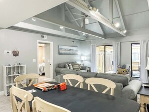 Un salon moderne comprenant un coin repas, des canapés gris, une table noire et un ventilateur de plafond.