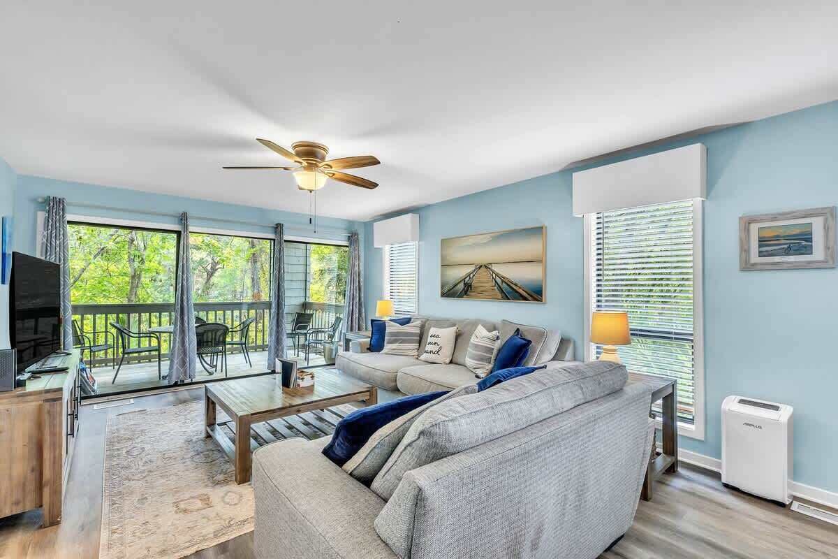 A living room with a sofa, coffee table, and TV, a balcony with outdoor furniture, and a ceiling fan.