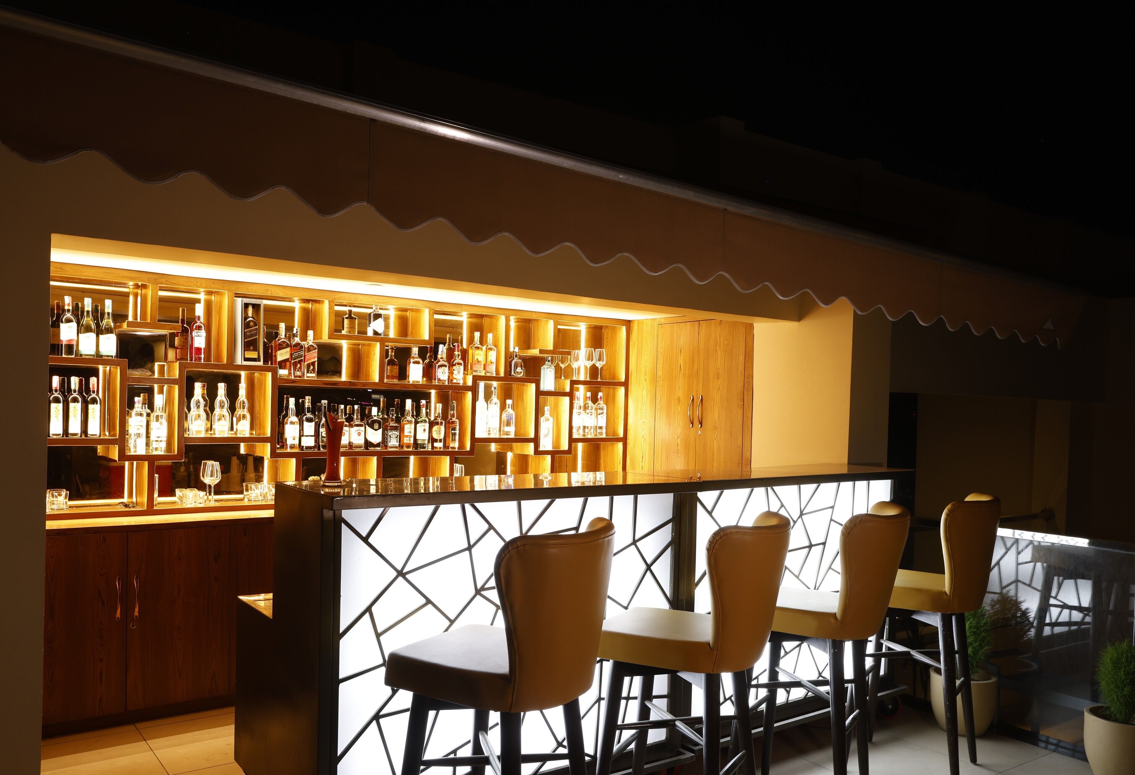 A bar area with wooden shelves stocked with bottles and glasses, a counter, and four yellow chairs.