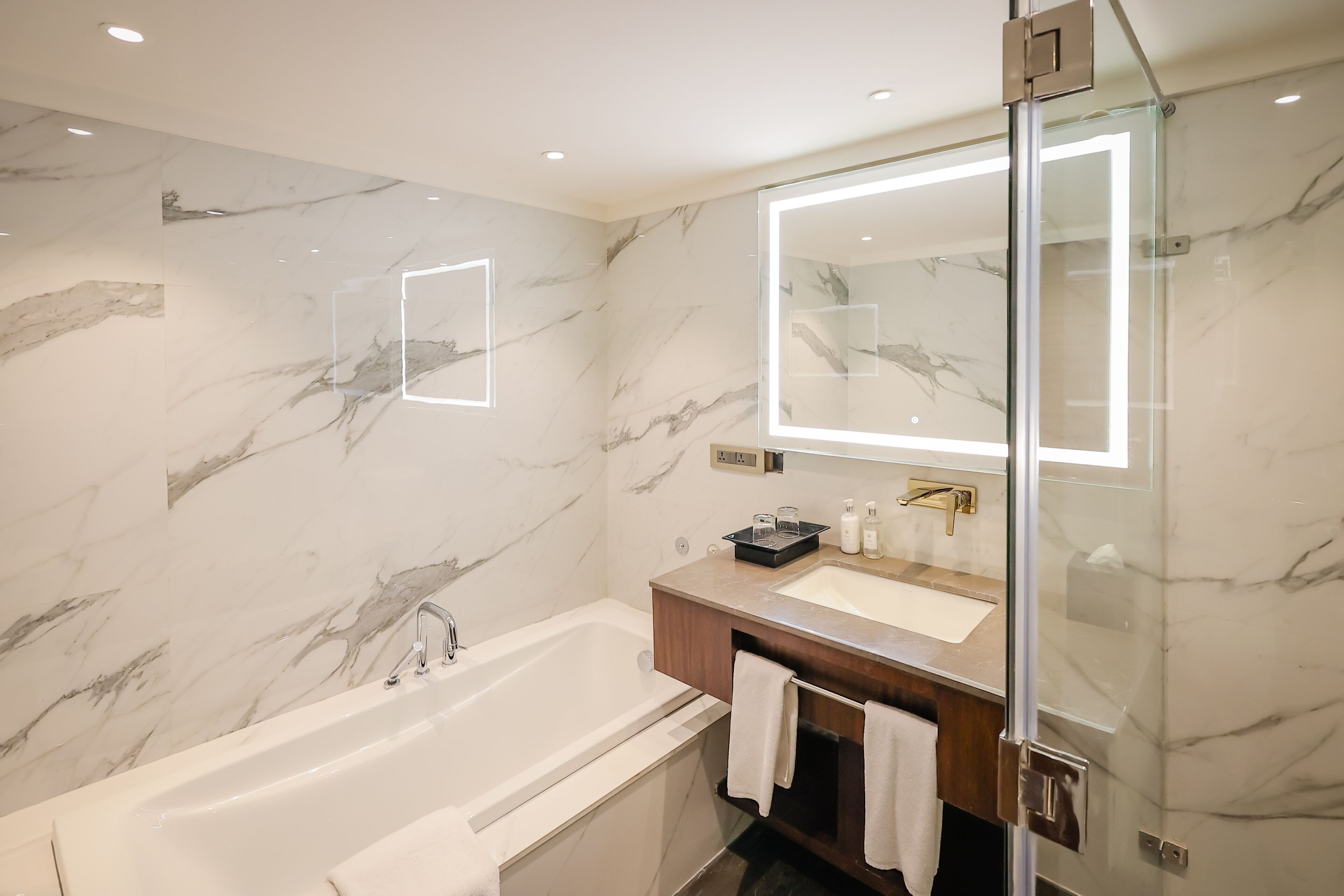 A modern bathroom with a marble wall, a large mirror, a bathtub, and a vanity with a sink and towel rack.