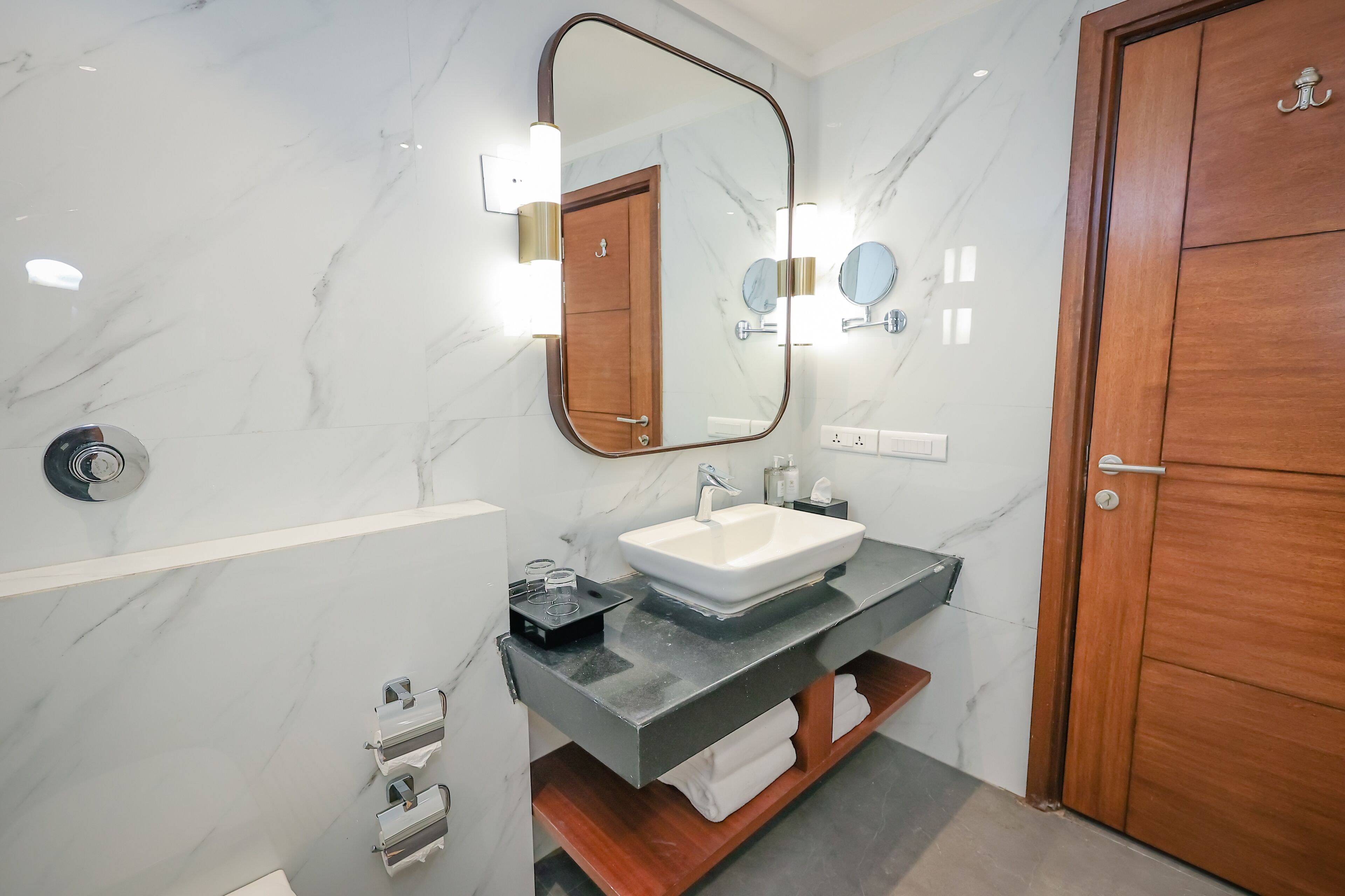 A modern bathroom with a marble wall, a large mirror, a sink, and a wooden door.
