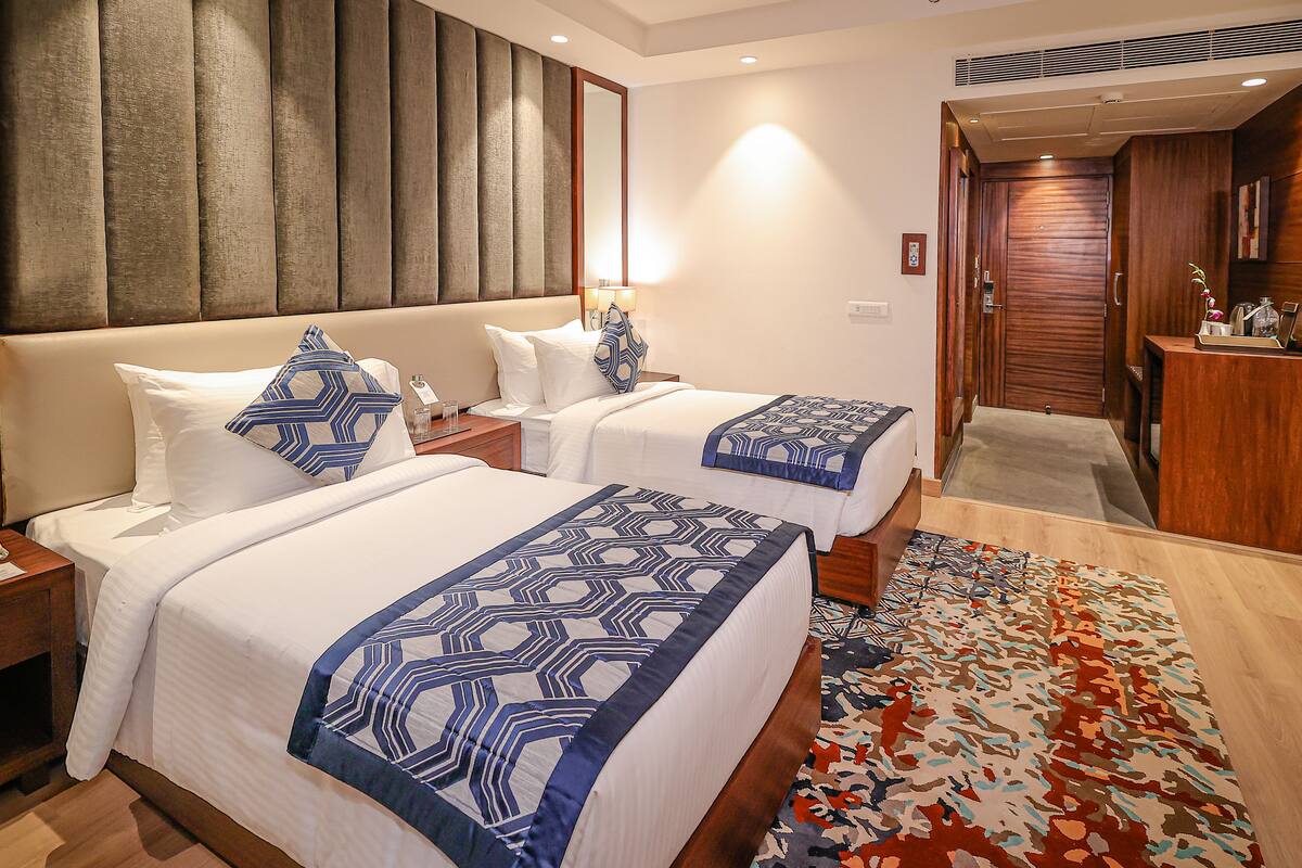 A hotel room with two beds, a wooden headboard, a nightstand, a bedside table, a lamp, a mirror, a wooden desk, and a patterned rug.