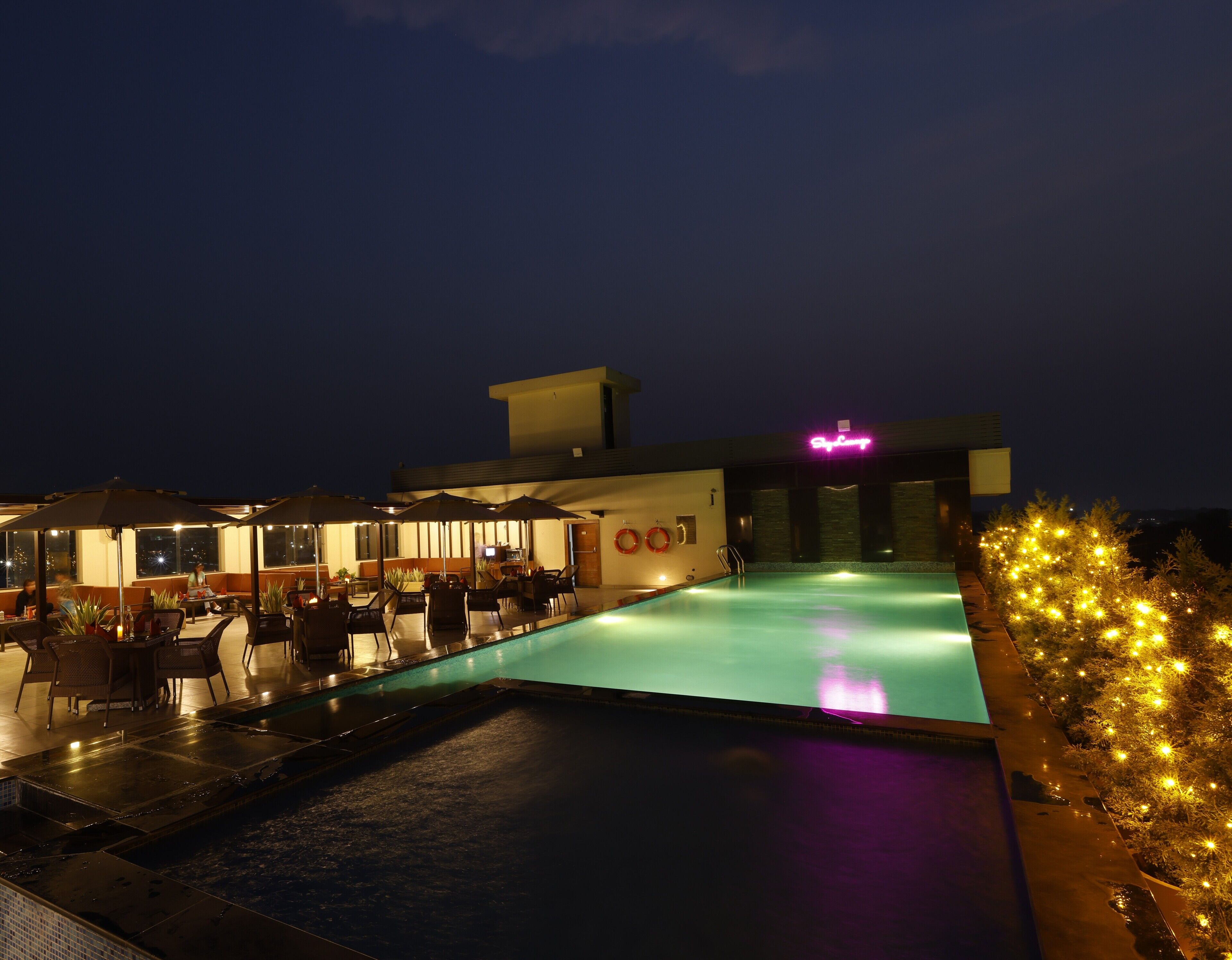 A rooftop pool area with lounge chairs and umbrellas, a neon sign, and a poolside bar.