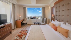 Ein Hotelzimmer mit einem großen Bett, einem Schreibtisch, einem Sessel, einem Fernseher und Blick auf die Stadt durch große Fenster.