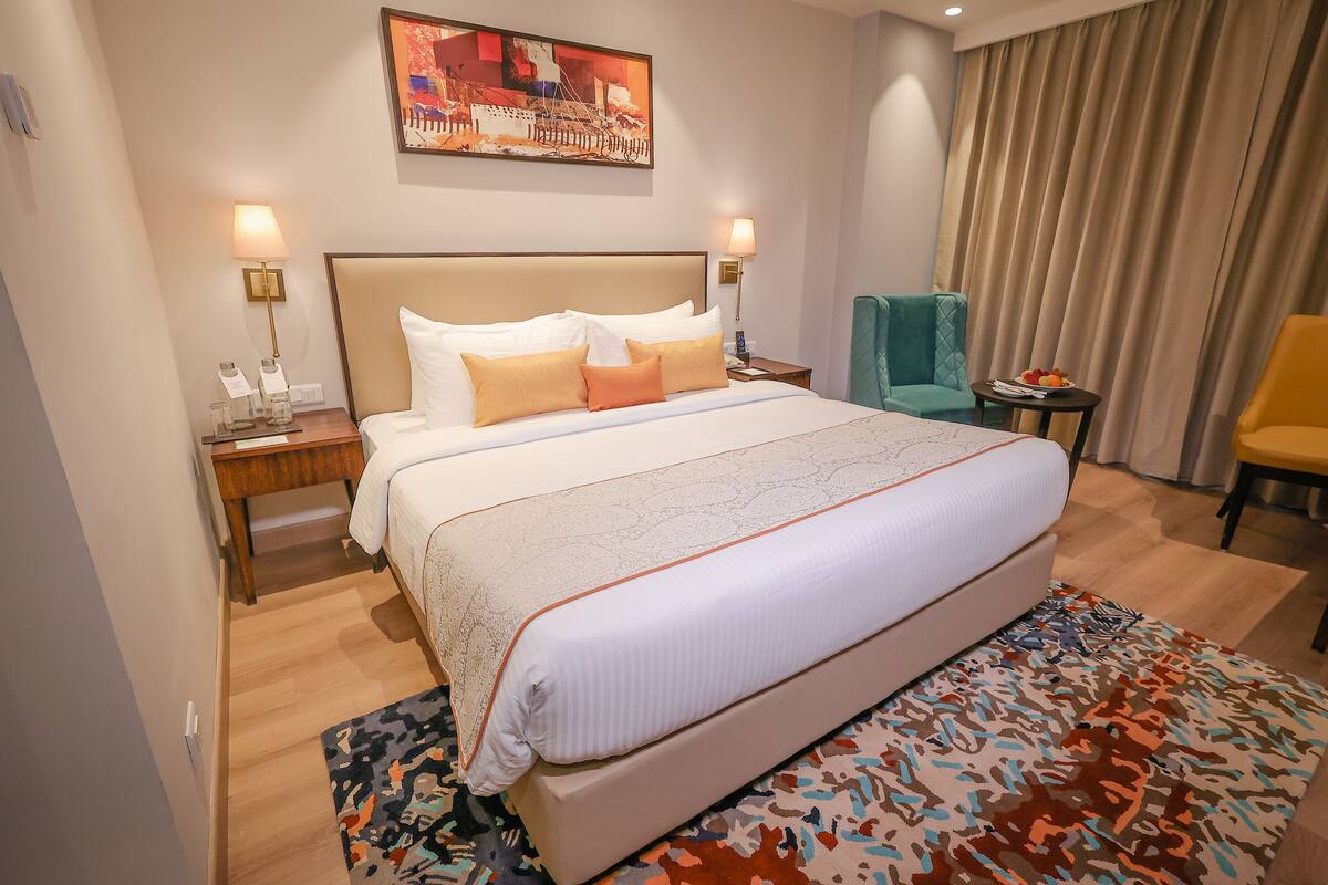 A hotel room with a large bed, two bedside tables with lamps, a chair, a small table with fruit, and a painting on the wall.