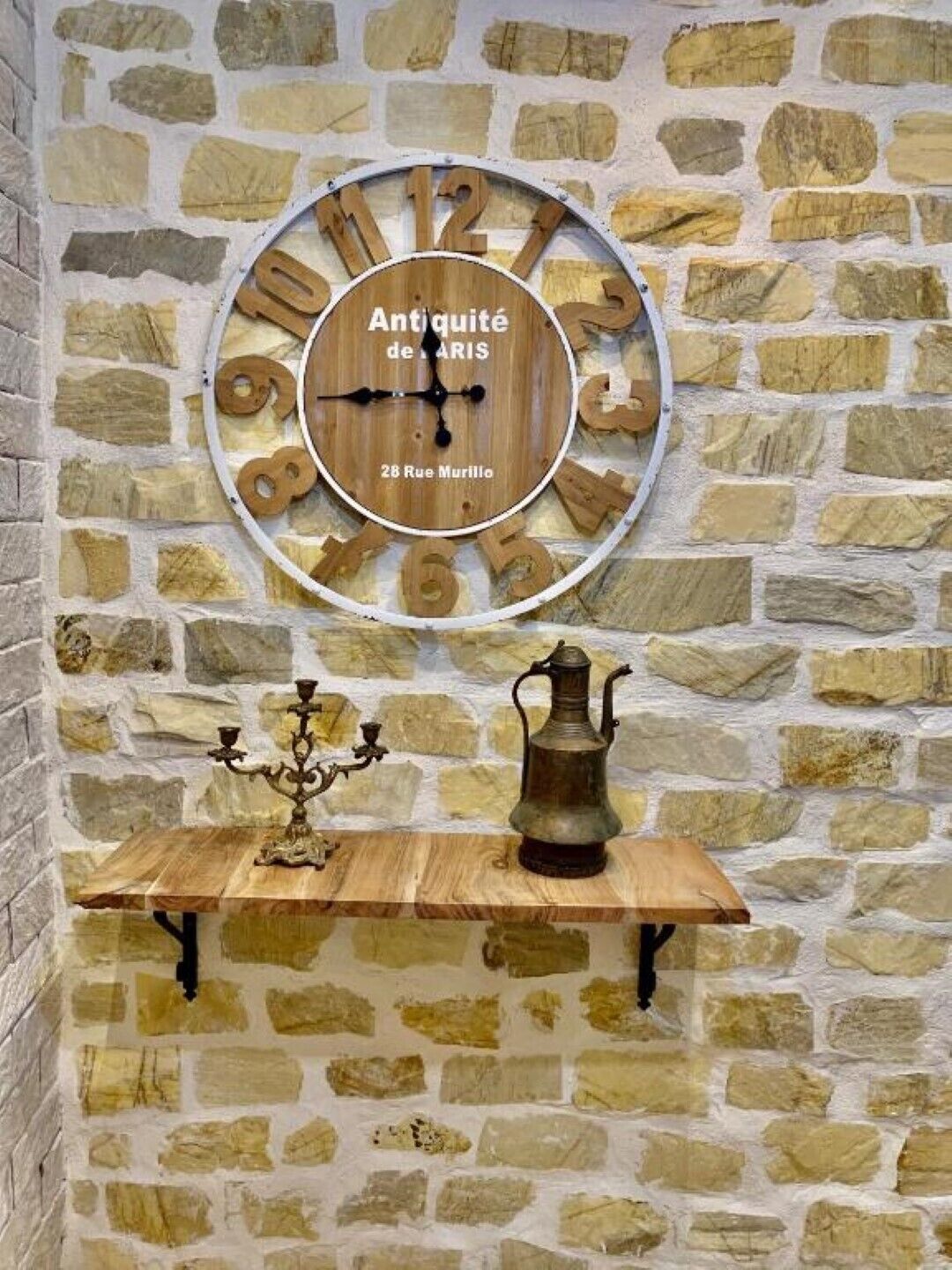 A stone wall with a wooden clock and a small table holding a candelabra and a metal pitcher.