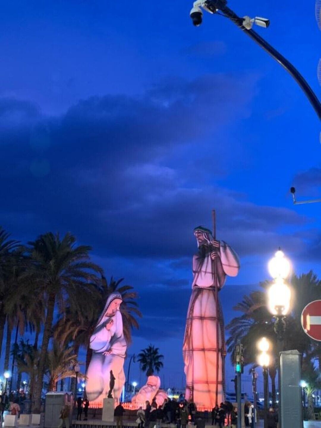 A nighttime view of a public square with illuminated statues and palm trees.
