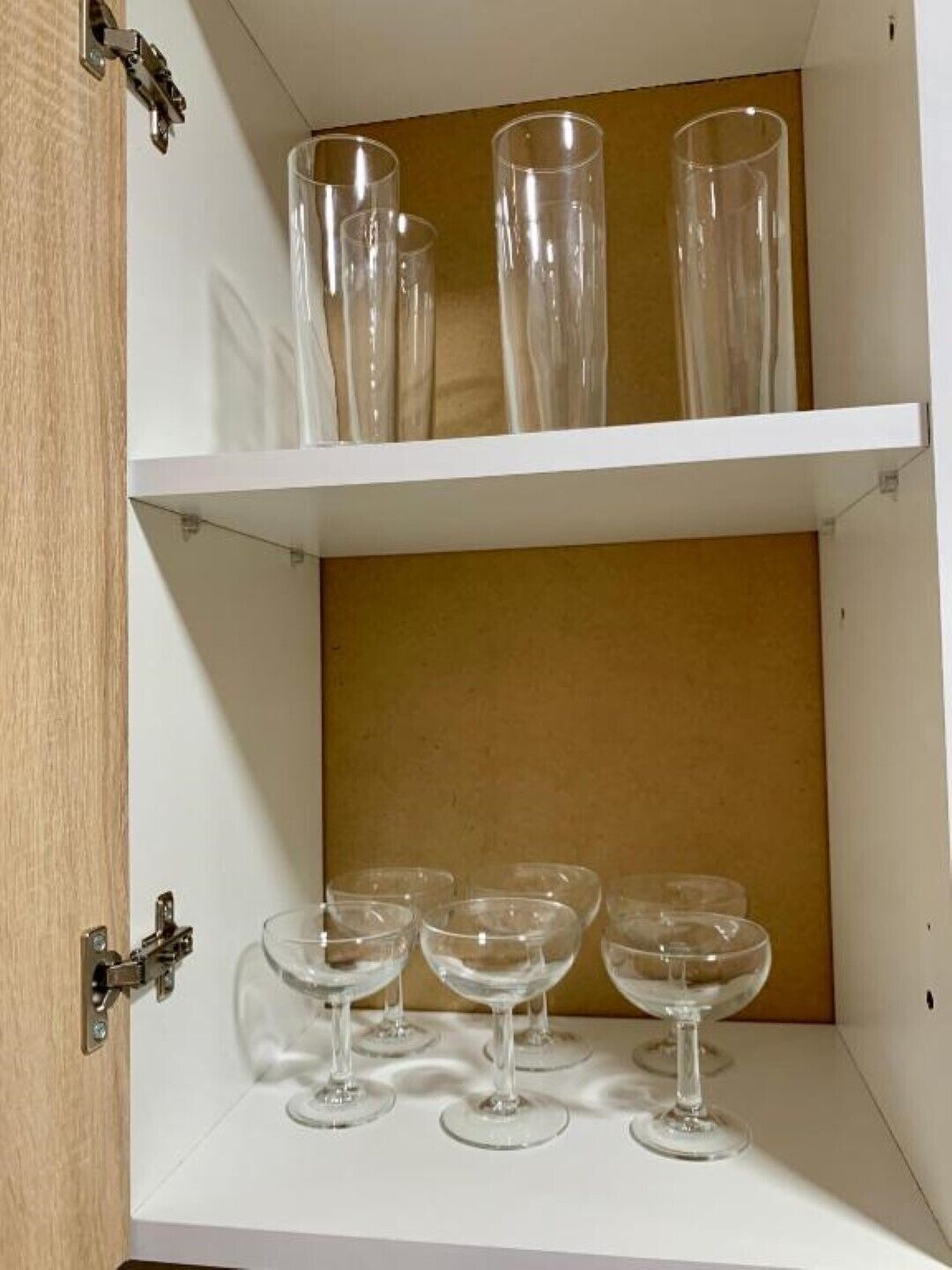 Un mueble blanco con dos estantes, uno con vasos altos y transparentes y el otro con vasos más bajos y transparentes.