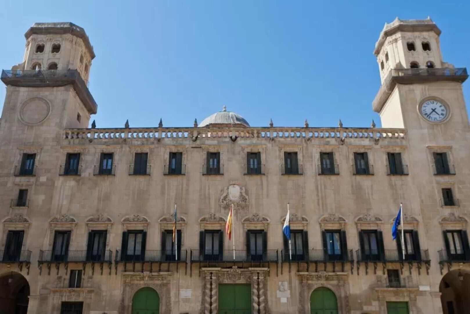 A large, historic building with two clock towers, a central dome, and multiple flags.