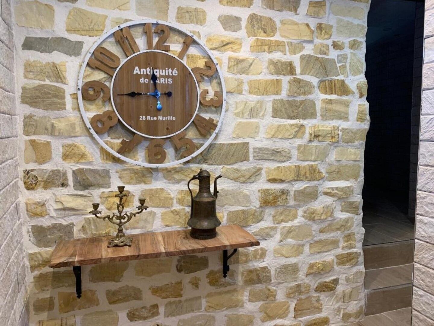 Un muro de piedra con un reloj de madera y una pequeña mesa con un candelabro y una jarrón.