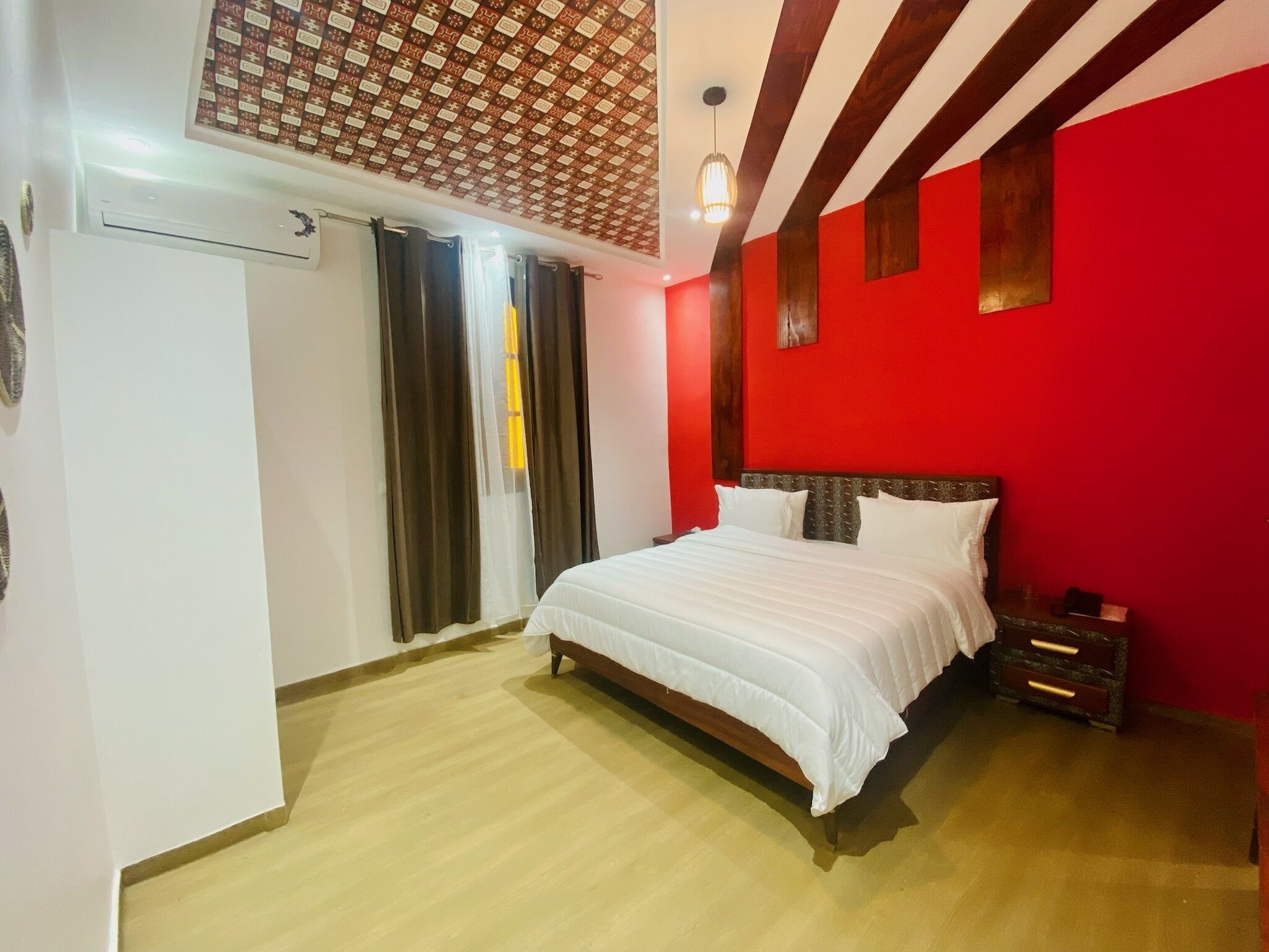 A bedroom with a bed, a nightstand, a red wall, a wooden floor, and a patterned ceiling.