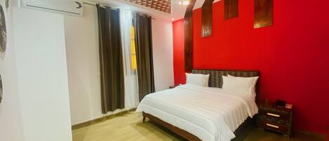 A bedroom with a bed, a nightstand, a red wall, a wooden floor, and a patterned ceiling.