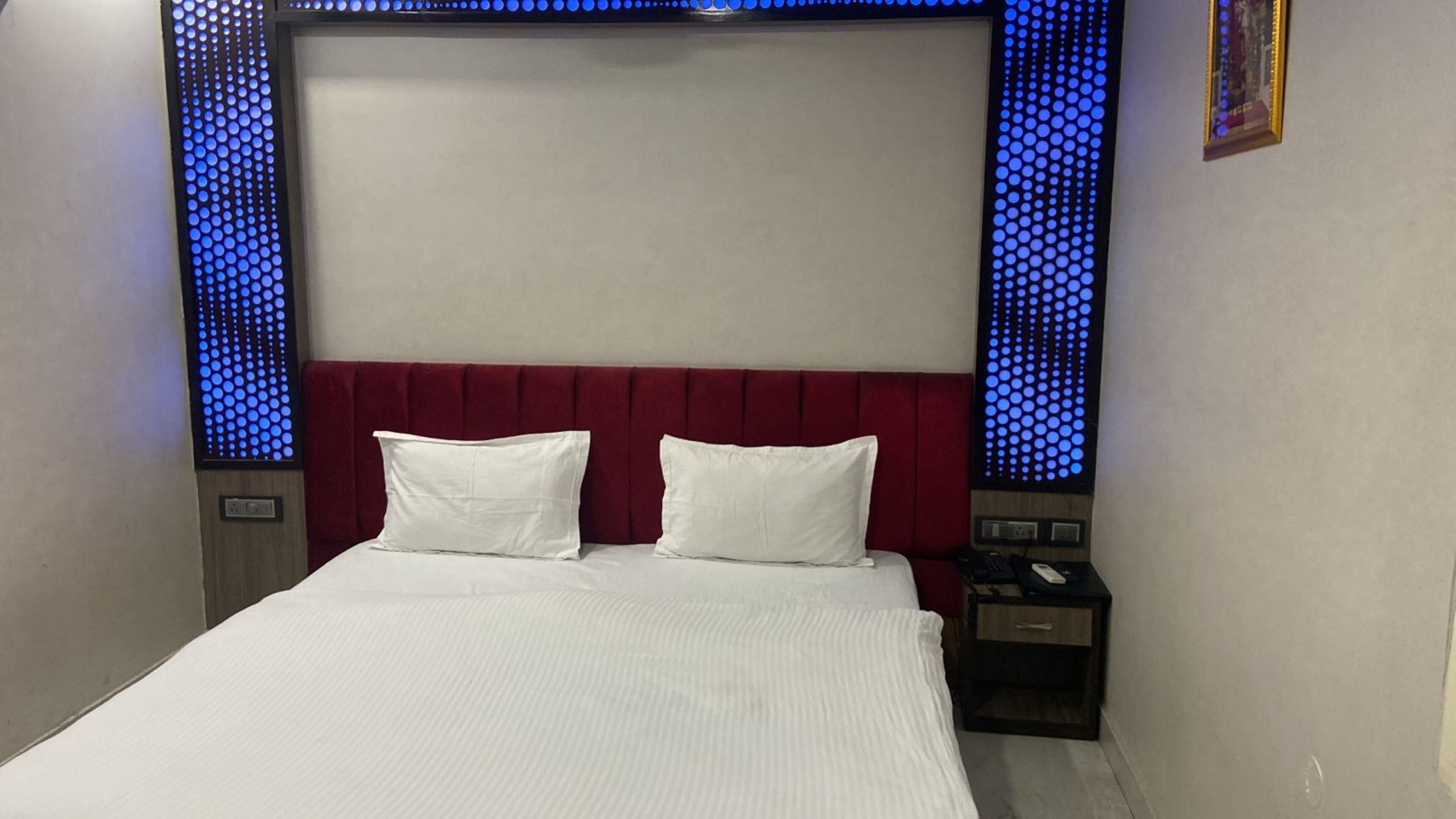 A hotel room with a bed, two pillows, a nightstand, a phone, and a wall with blue lighting.