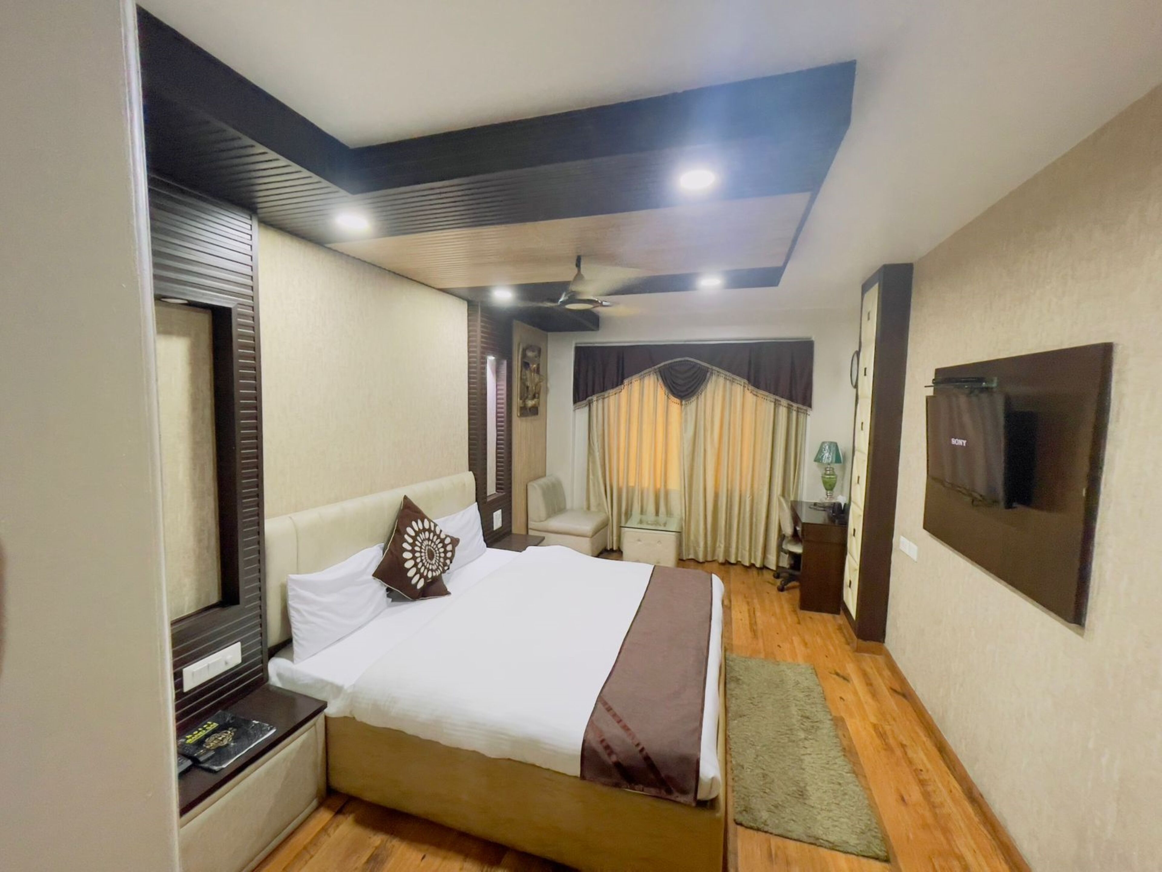 A hotel room with a bed, a nightstand, a desk, a chair, a TV mounted on the wall, and a ceiling fan.