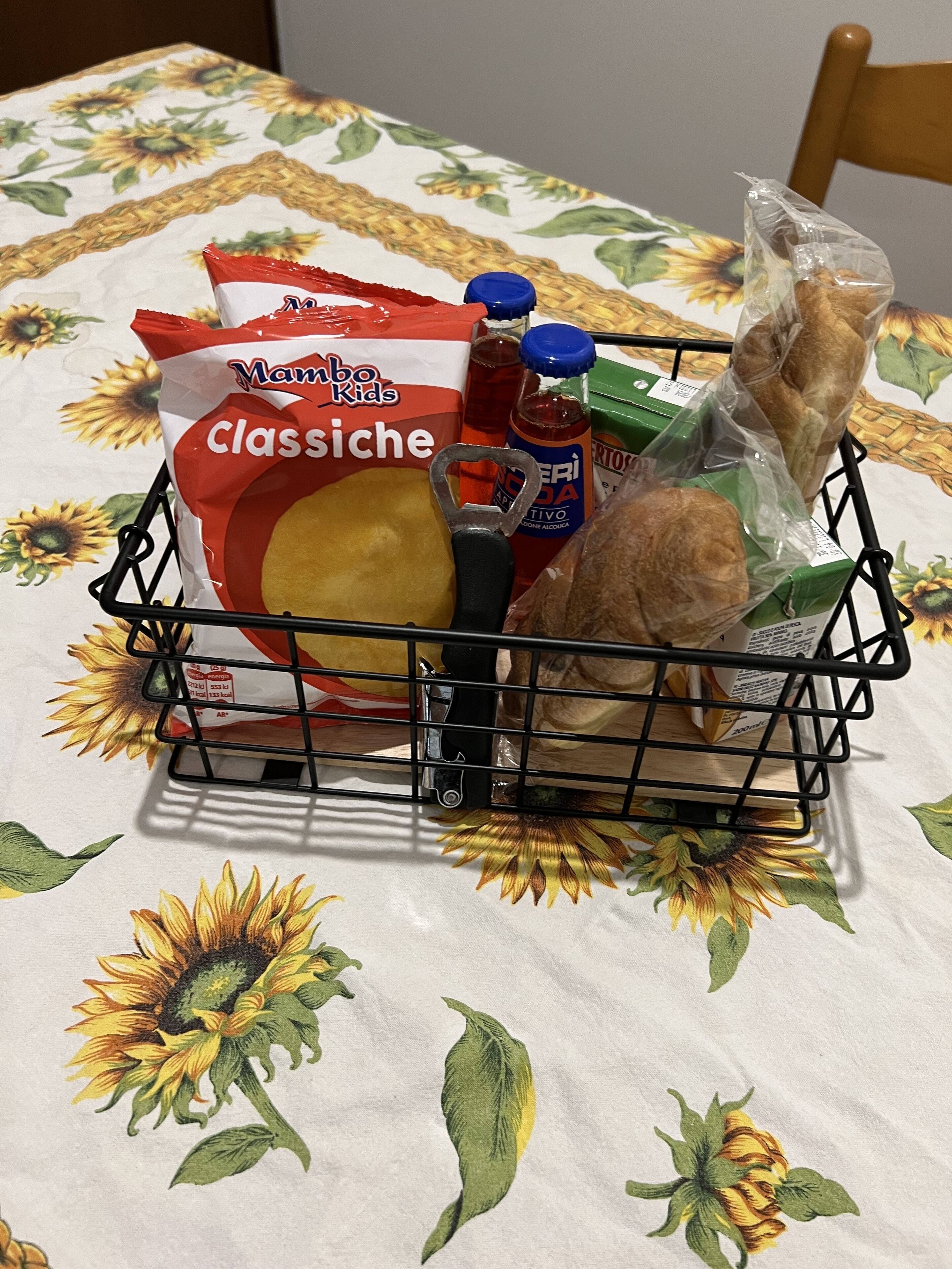 A shopping basket containing snacks and drinks on a table with a sunflower tablecloth.