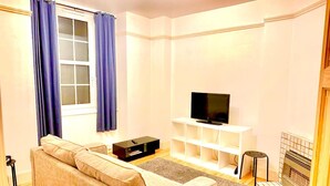 A living room with a sofa, a TV on a white stand, a fireplace, and a window with curtains. - Immaculate One Bed in Tower Bridge Luxurious Taste (London)