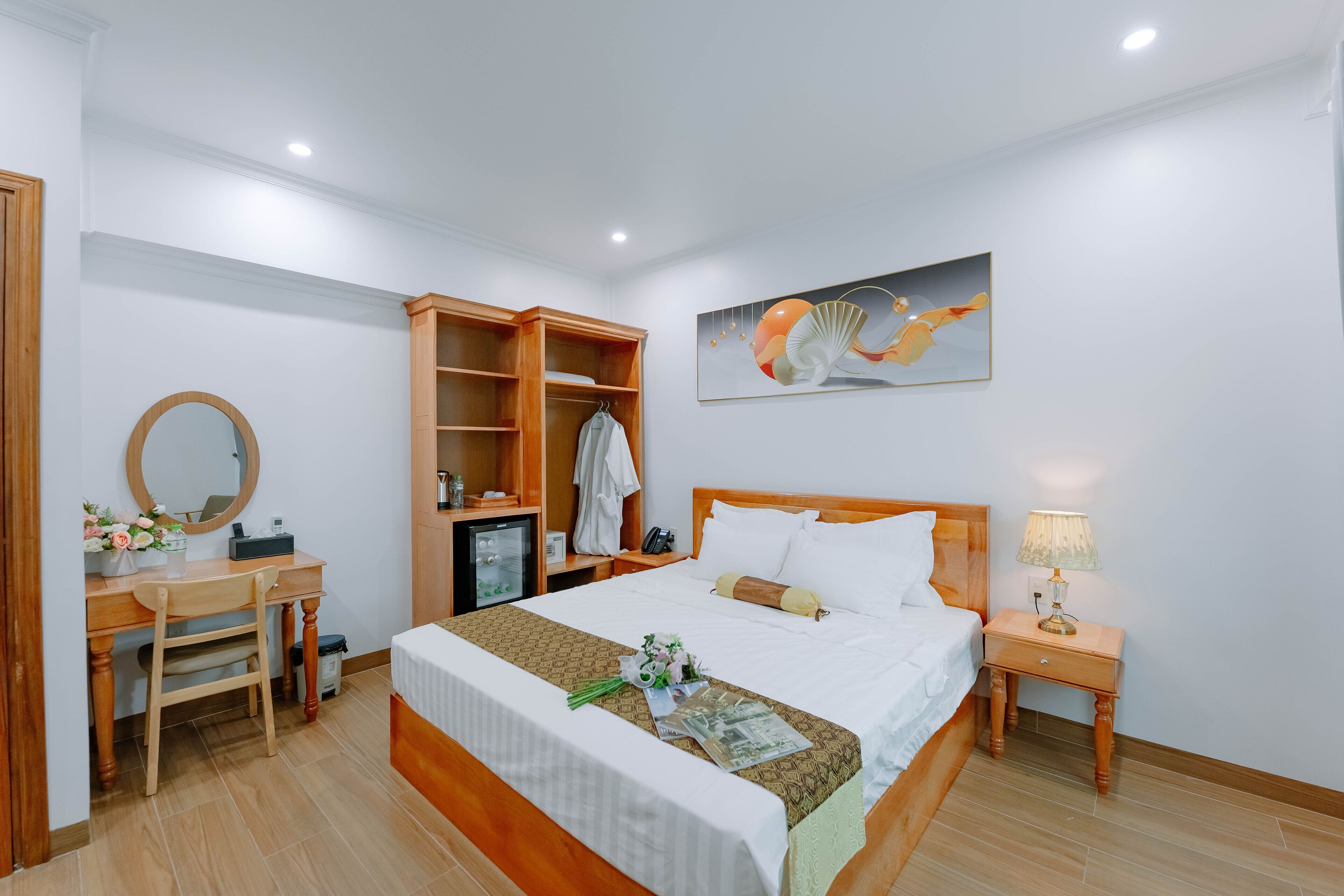 A hotel room with a bed, a desk with a chair, a wardrobe, a bedside table with a lamp, and a painting on the wall.