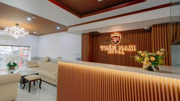 A hotel reception area with a wooden reception desk, a chandelier, a sofa, and a vase of flowers. - Tuan Nam Hotel (Hai Phong)
