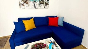 A blue sectional sofa with colorful cushions, a white coffee table with magazines and a remote control, and a beach-themed painting on the wall. - Suite del Ayuntamiento (Alicante (Alacant))