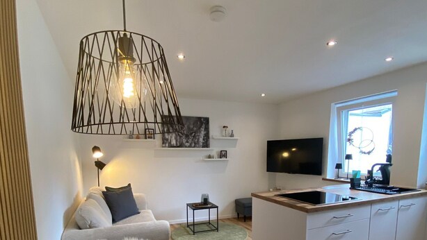 Una cocina moderna con una isla central, una zona de comedor con una mesa blanca y un salón con sofá y televisión.